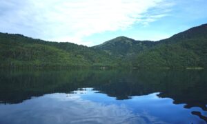 A tranquil mountain lake reflects lush green forest-covered hills and a bright cloud-filled sky, creating a peaceful scene of natural beauty and symmetry.