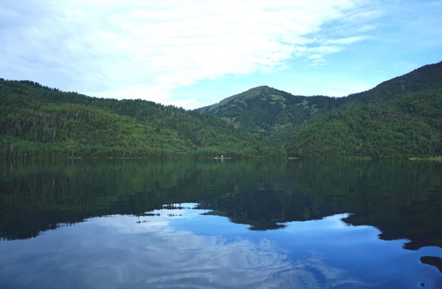 A tranquil mountain lake reflects lush green forest-covered hills and a bright cloud-filled sky, creating a peaceful scene of natural beauty and symmetry.