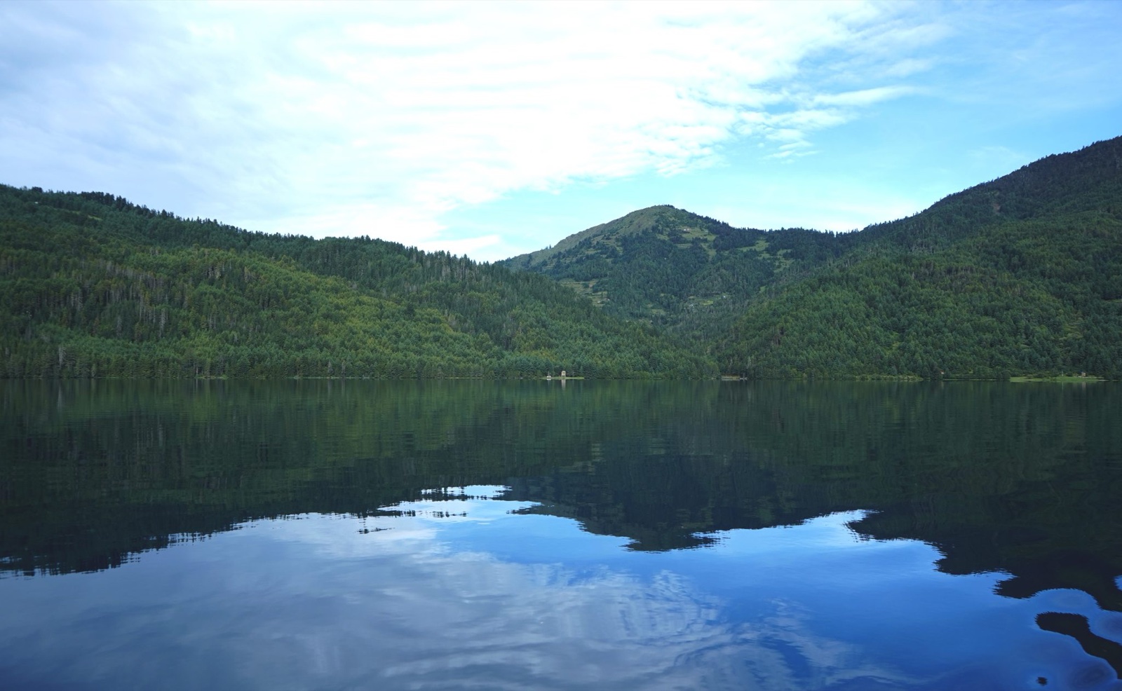 A tranquil mountain lake reflects lush green forest-covered hills and a bright cloud-filled sky, creating a peaceful scene of natural beauty and symmetry.