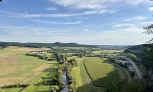 A sweeping rural landscape of rolling hills, patchwork fields, and a winding tree-lined river under a clear blue sky. A cluster of buildings and a curving road in the foreground hint at a nearby village amid the peaceful countryside.