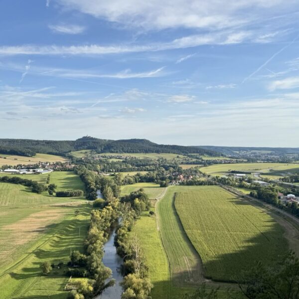 A sweeping rural landscape of rolling hills, patchwork fields, and a winding tree-lined river under a clear blue sky. A cluster of buildings and a curving road in the foreground hint at a nearby village amid the peaceful countryside.