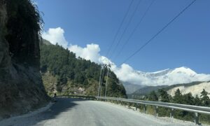 A winding mountain road with a gravel surface, flanked by steep rocky cliffs on the left and dense green pine trees on the right.