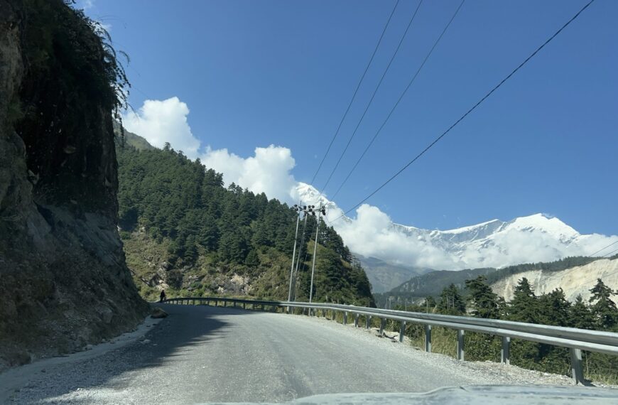 A winding mountain road with a gravel surface, flanked by steep rocky cliffs on the left and dense green pine trees on the right.