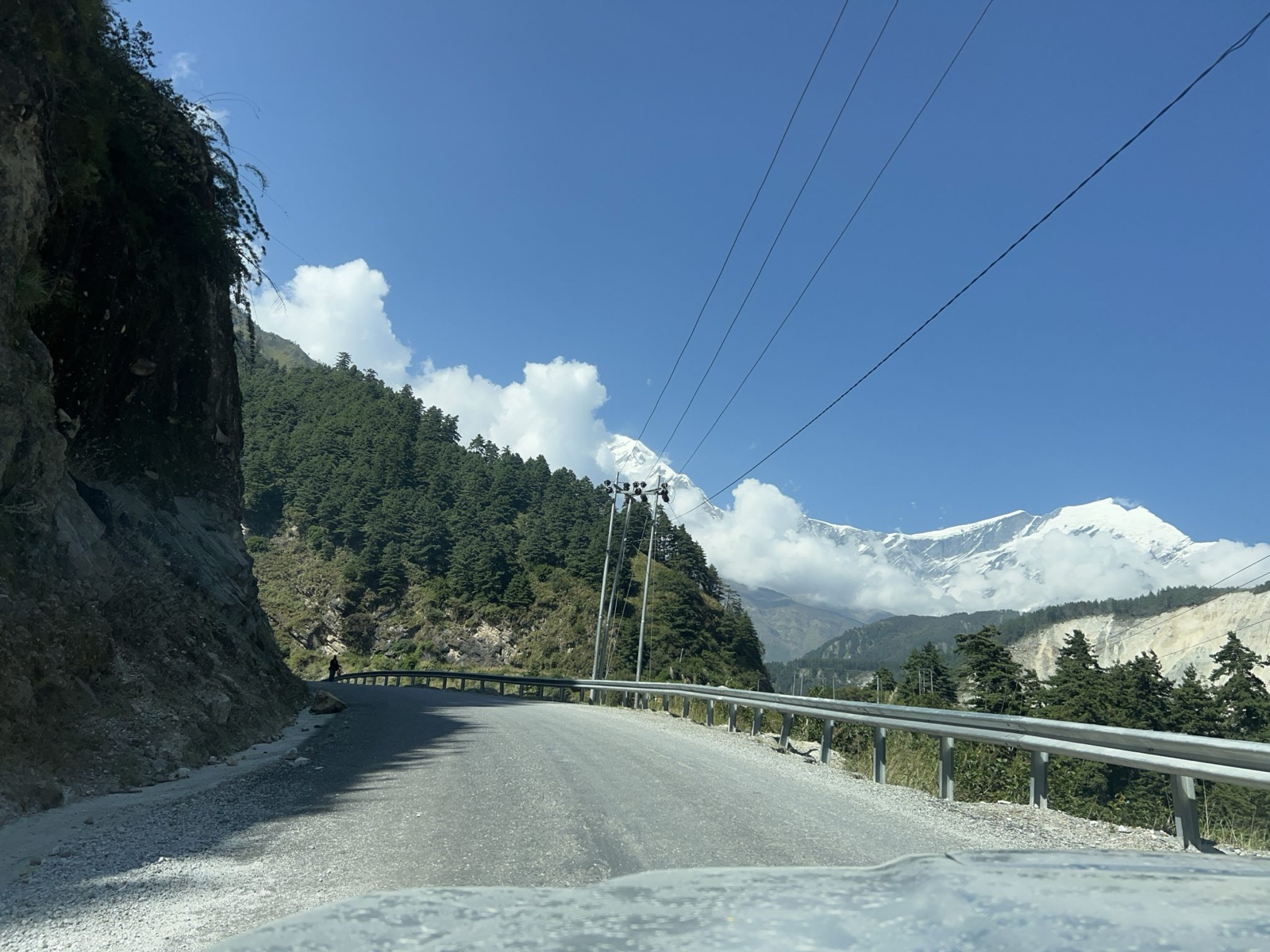 A winding mountain road with a gravel surface, flanked by steep rocky cliffs on the left and dense green pine trees on the right.