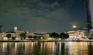 A serene night view of Singapore’s waterfront, showcasing the beautifully illuminated Asian Civilisations Museum and the iconic Fullerton Hotel. The calm waters reflect the warm lights from the historic buildings and surrounding structures, while the full moon peeks through scattered clouds, adding a touch of tranquility to the vibrant cityscape.