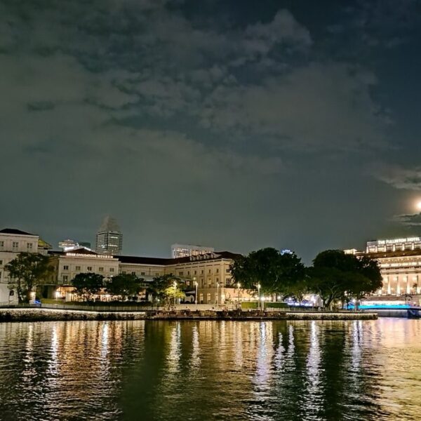 A serene night view of Singapore’s waterfront, showcasing the beautifully illuminated Asian Civilisations Museum and the iconic Fullerton Hotel. The calm waters reflect the warm lights from the historic buildings and surrounding structures, while the full moon peeks through scattered clouds, adding a touch of tranquility to the vibrant cityscape.