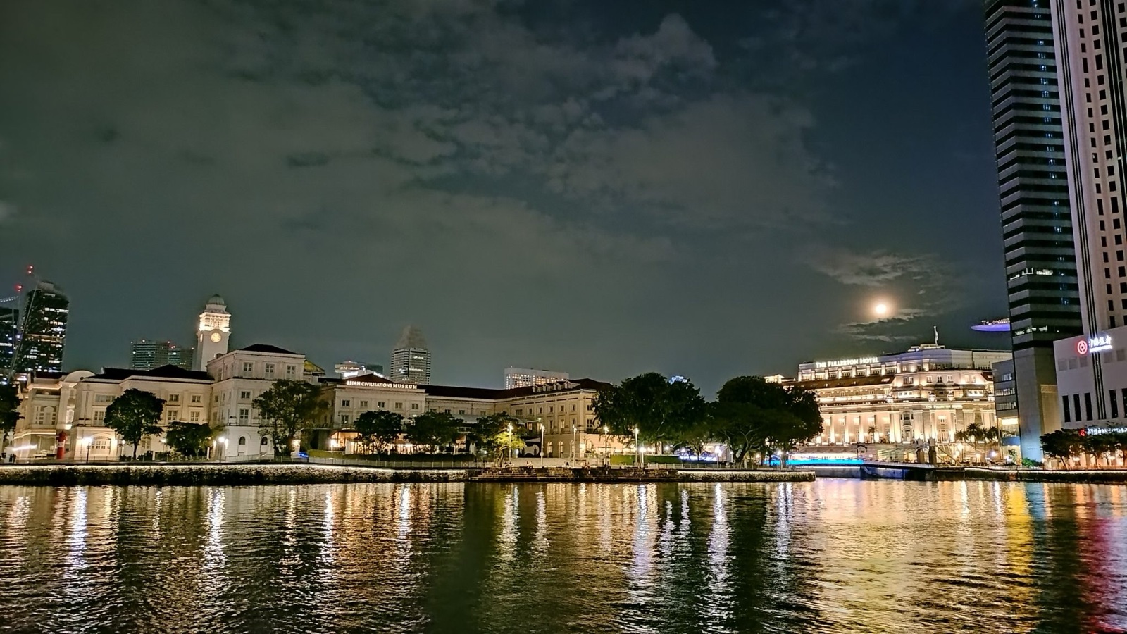 A serene night view of Singapore’s waterfront, showcasing the beautifully illuminated Asian Civilisations Museum and the iconic Fullerton Hotel. The calm waters reflect the warm lights from the historic buildings and surrounding structures, while the full moon peeks through scattered clouds, adding a touch of tranquility to the vibrant cityscape.